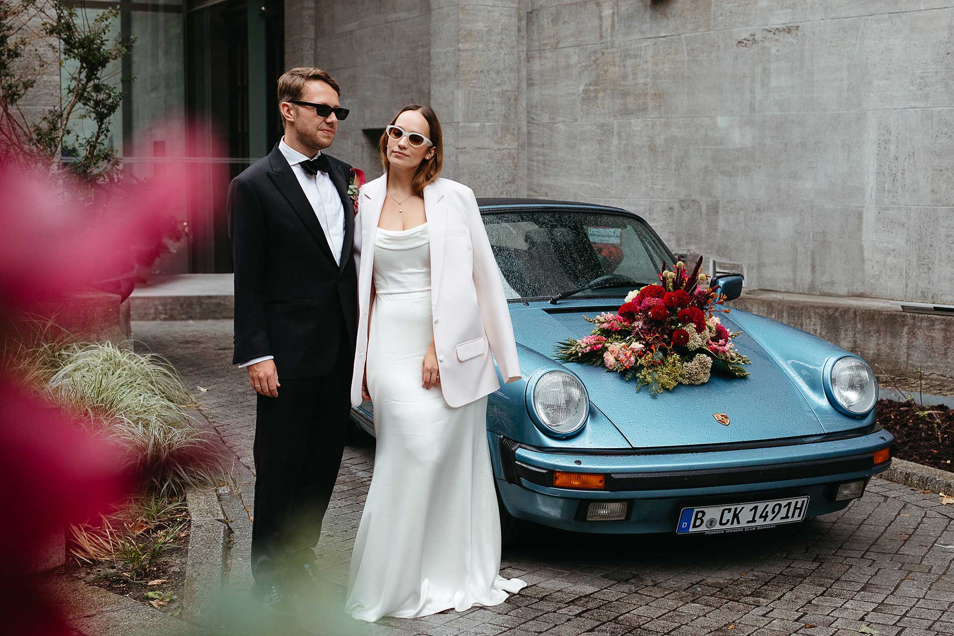 Emotionale Hochzeitsfotos: Braut und Bräutigam beide mit Sonnenbrille vor ihrem Hochzeitsauto, einem Porsche Oldtimer. Bild von Hochzeitsfotografin Laura-Sophie Jung, Berlin.