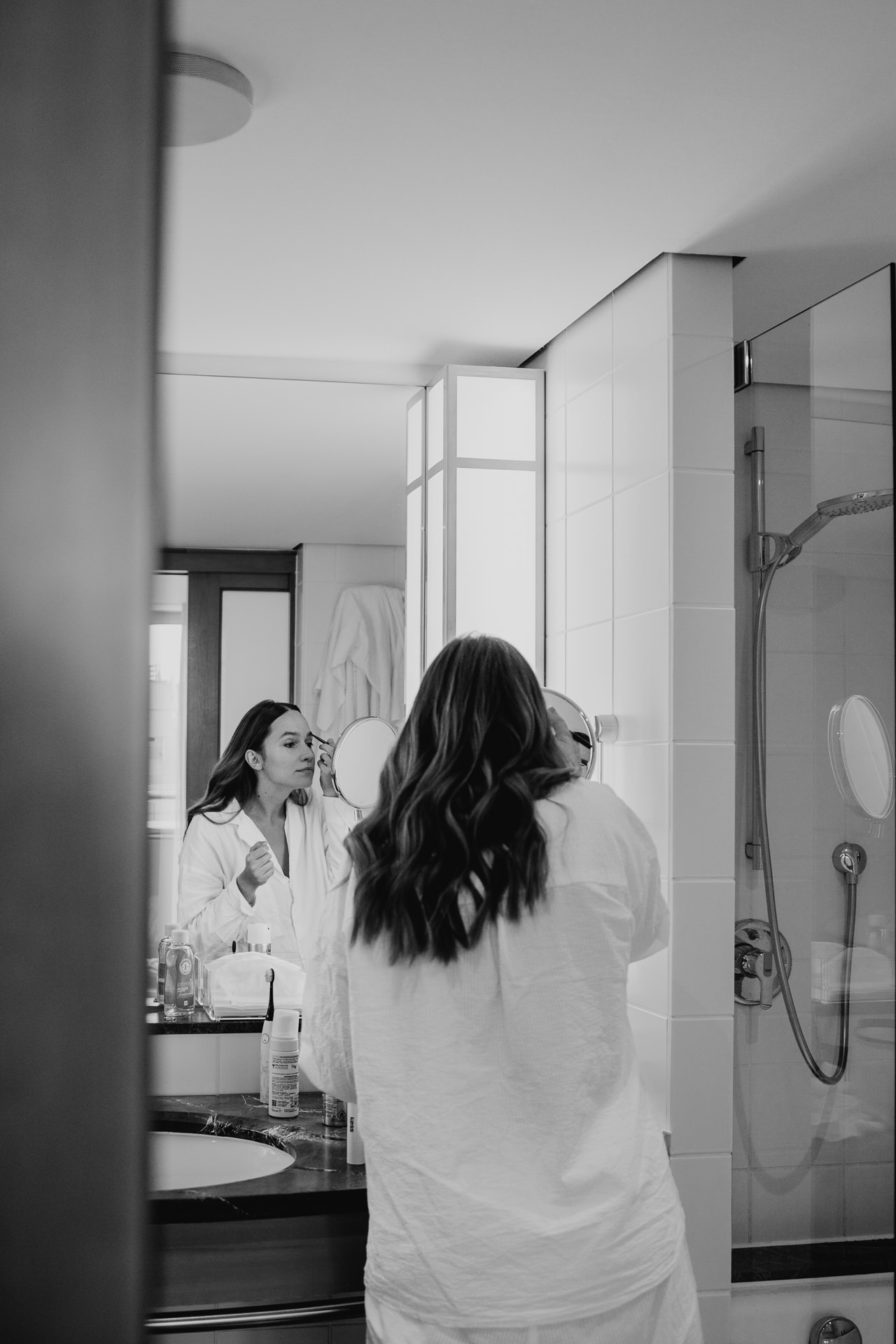 Getting Ready für die Hochzeit: Braut steht vor dem Spiegel und schminkt sich. Aufnahme in schwarz-weiß, fotografiert von Laura-Sophie Jung.