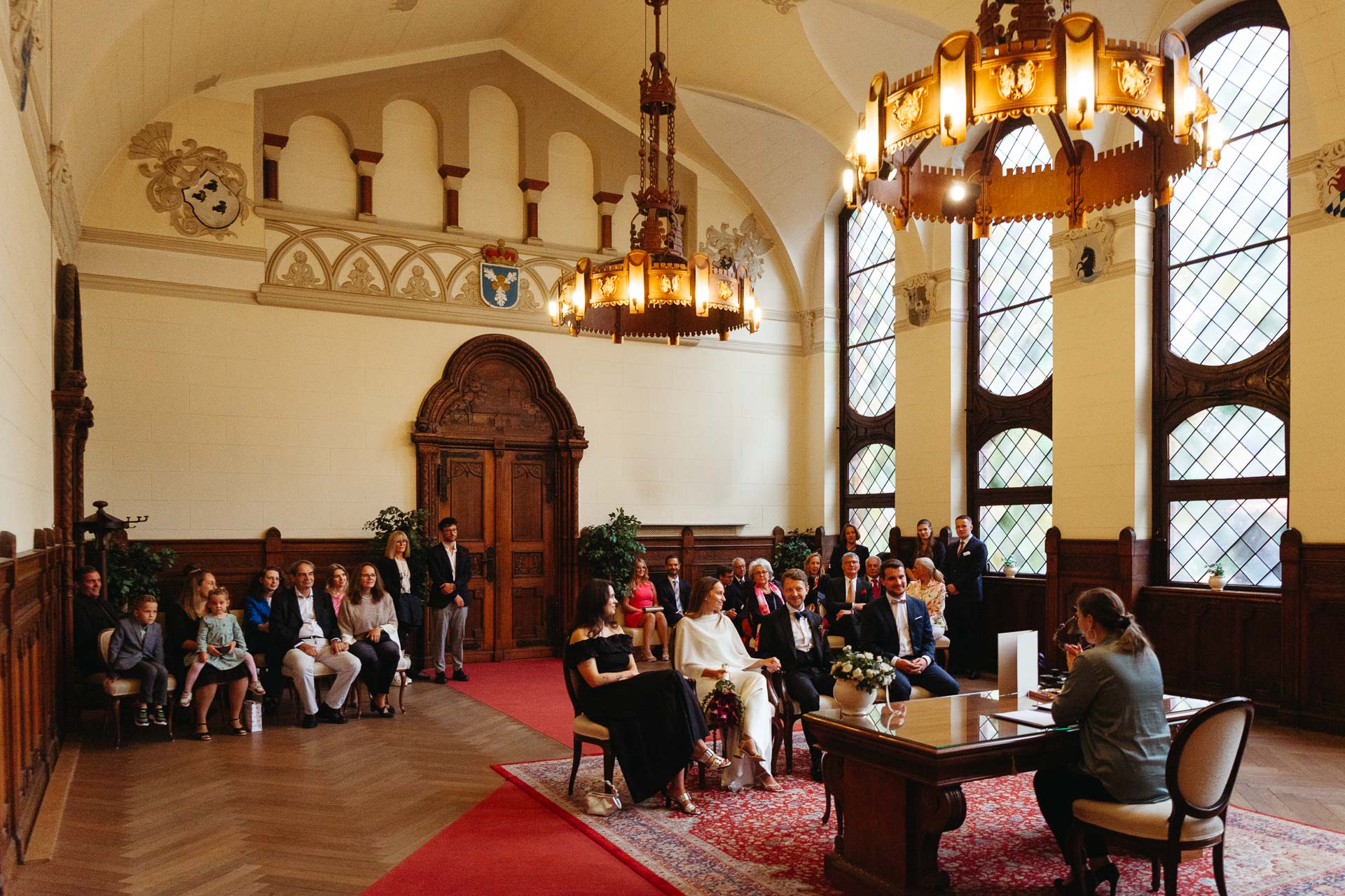 Standesamtliche Trauung im Rathaus Schmargendorf: Braut, Bräutigam, Trauzeuge und Trauzeugin sitzen im Trausaal vor der Standesbeamtin, Gäste im Hintergrund. Foto von Laura-Sophie Jung.