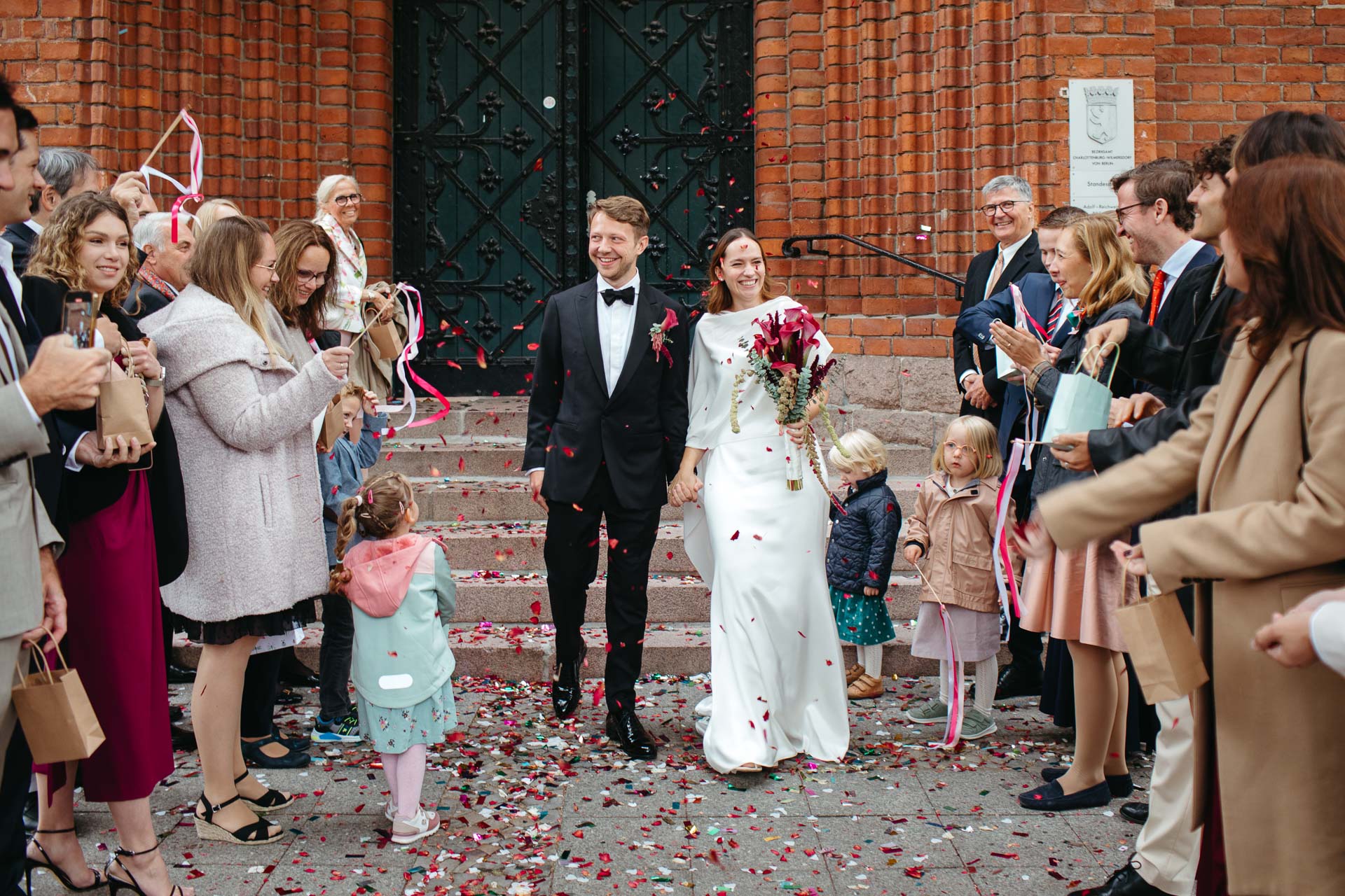 Emotionales Hochzeitsfoto: Auszug nach der Trauung. Glückliches Brautpaar kommt aus dem Standesamt, rechts und links Hochzeitsgäste und überall Konfetti. Foto von Laura-Sophie Jung.