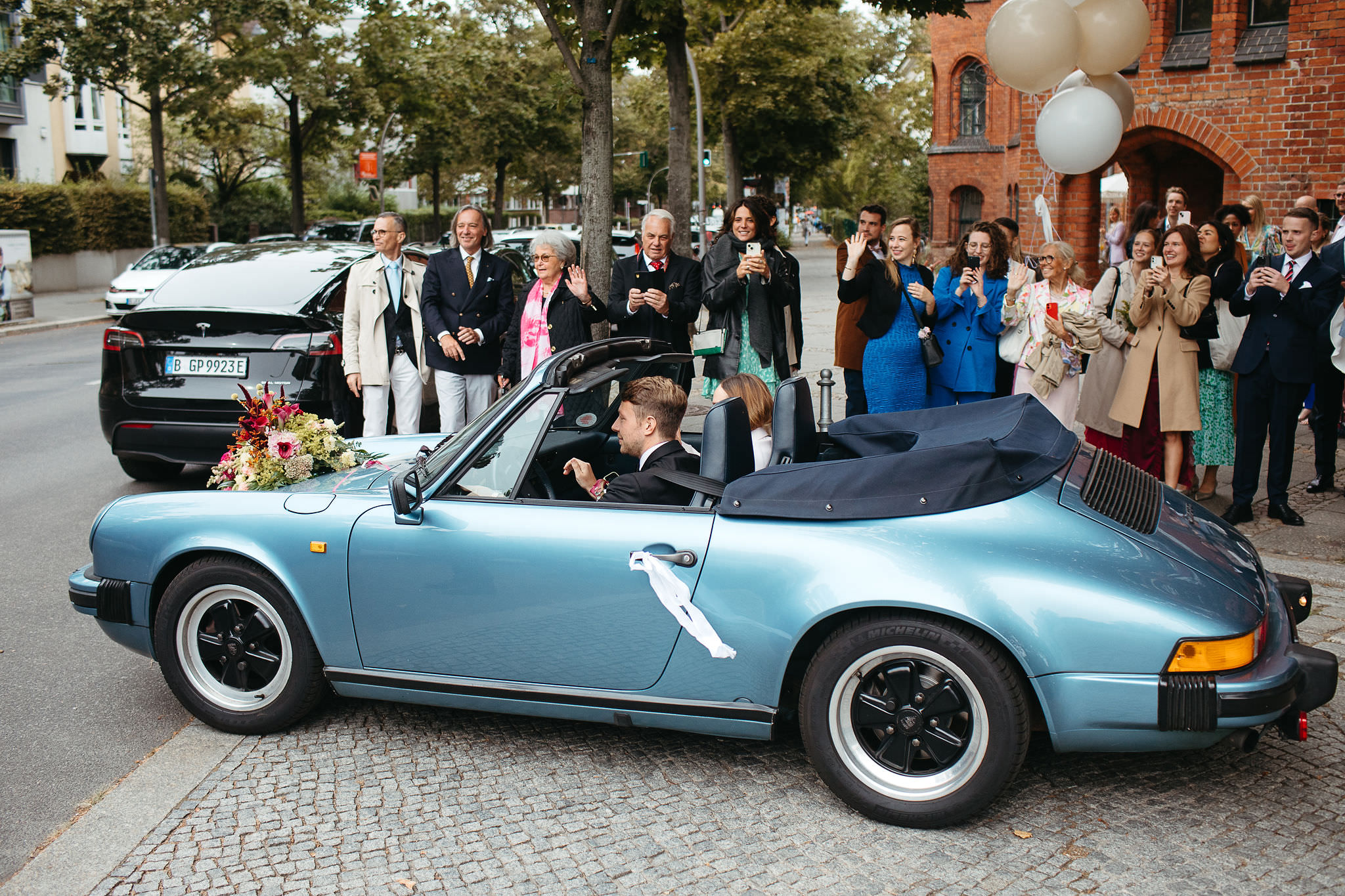 Vom Standesamt in die Hochzeitslocation zur Feier: Brautpaar im Hochzeitsauto und winkende Gäste. Foto von Laura-Sophie Jung.