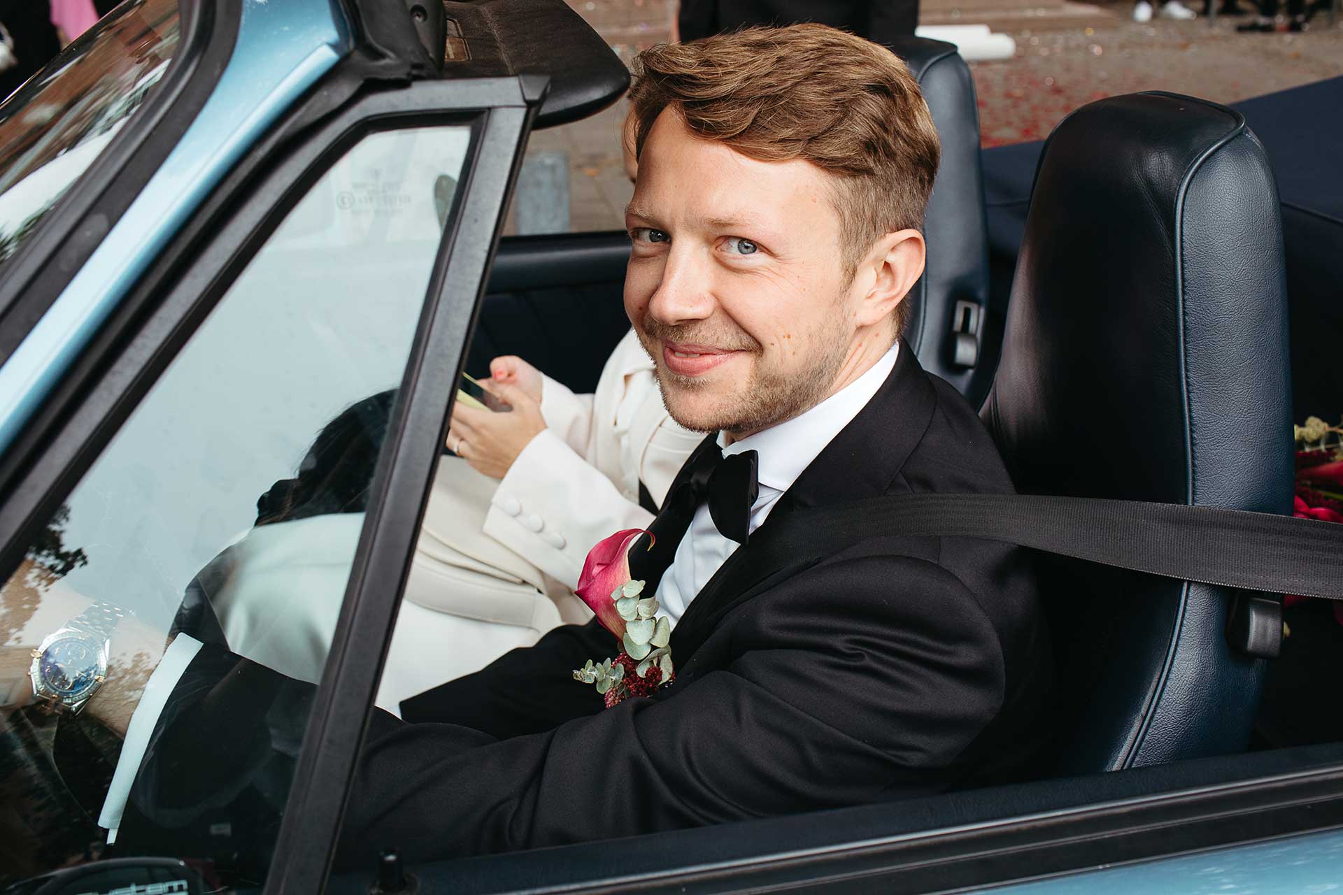 Brautpaar im Hochzeitsauto auf dem Weg zur Hochzeitsfeier. Bräutigam lächelt glücklich in die Kamera. Fotografiert von Laura-Sophie Jung.