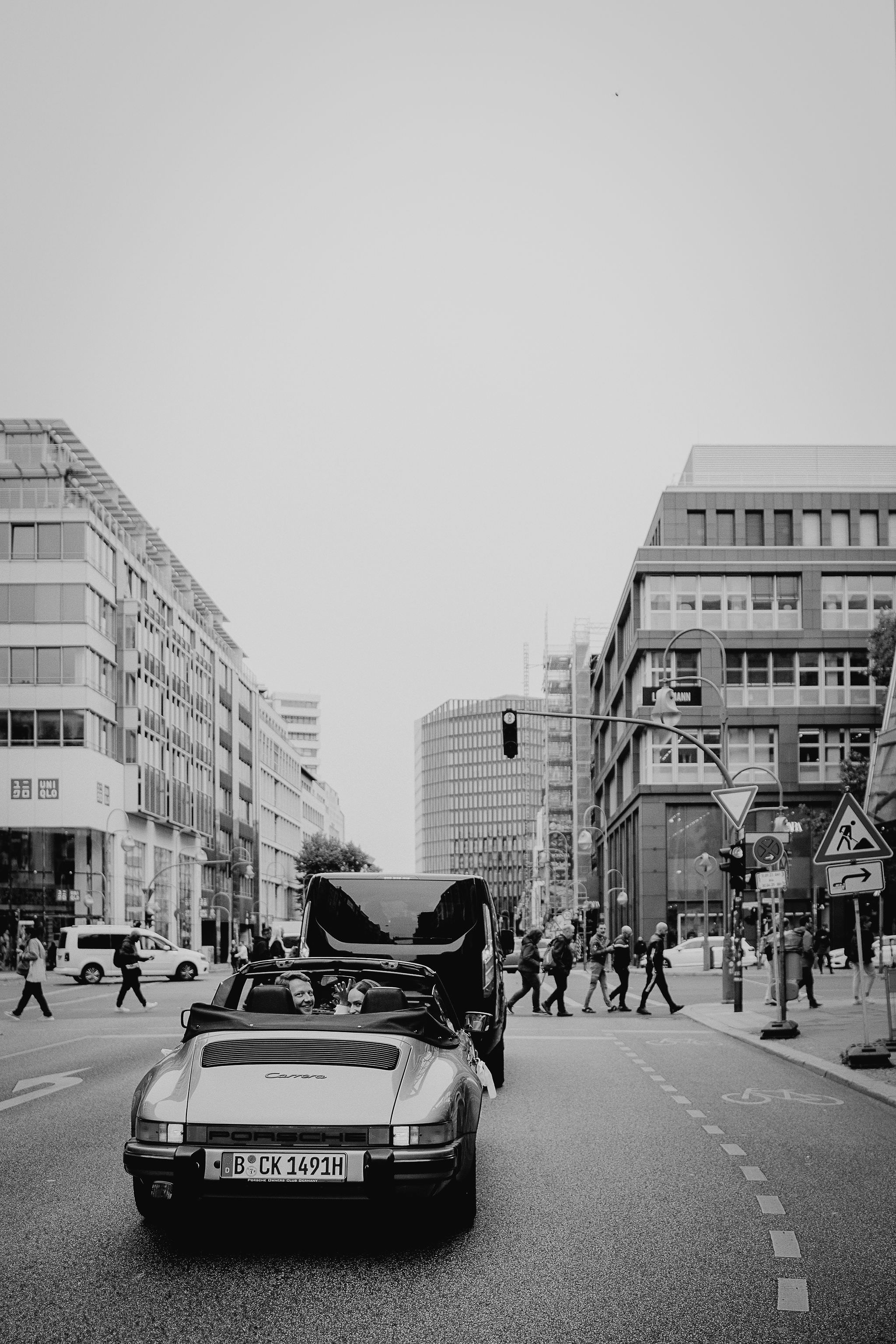 Brautpaar auf dem Weg zur Hochzeitsfeier winkt aus dem Hochzeitsauto - ein Porsche Cabrio Oldtimer. Fotografiert von Laura-Sophie Jung.