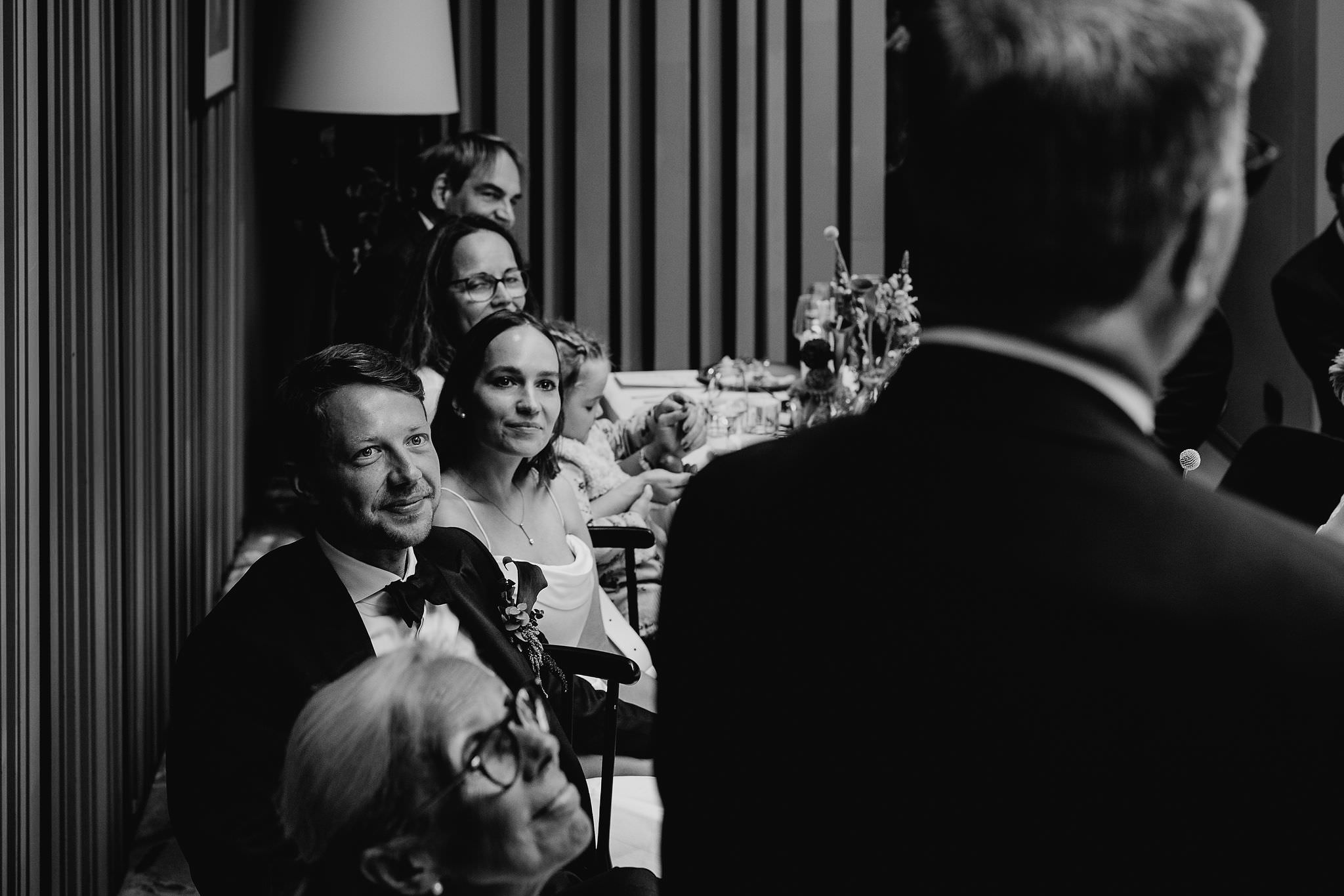 Hochzeitsfeier: Braut, Bräutigam und Gäste am Tisch, ein Hochzeitsgast hält eine Rede emotionale Rede. Foto in schwarz-weiß von Laura-Sophie Jung.