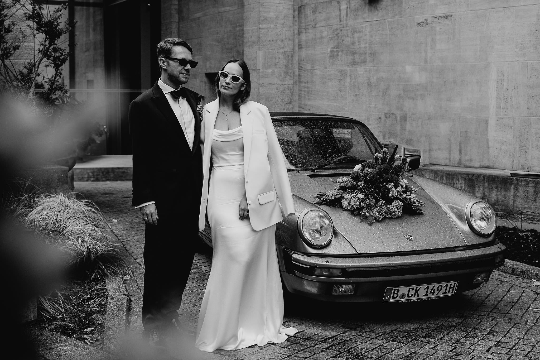Hochzeitsshooting: Brautpaar post vor dem Hochzeitsauto. Bild in schwarz-weiß von Laura-Sophie Jung, Hochzeitsfotografin Berlin.