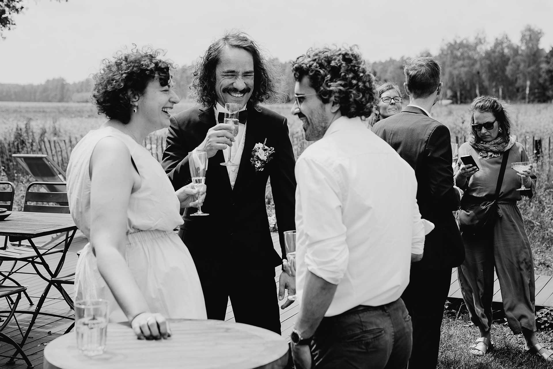 Hochzeit auf dem Land in Brandenburg: Bräutigam mit Gästen beim Sektempfang, fröhlich lachend. Foto von Laura-Sophie Jung.