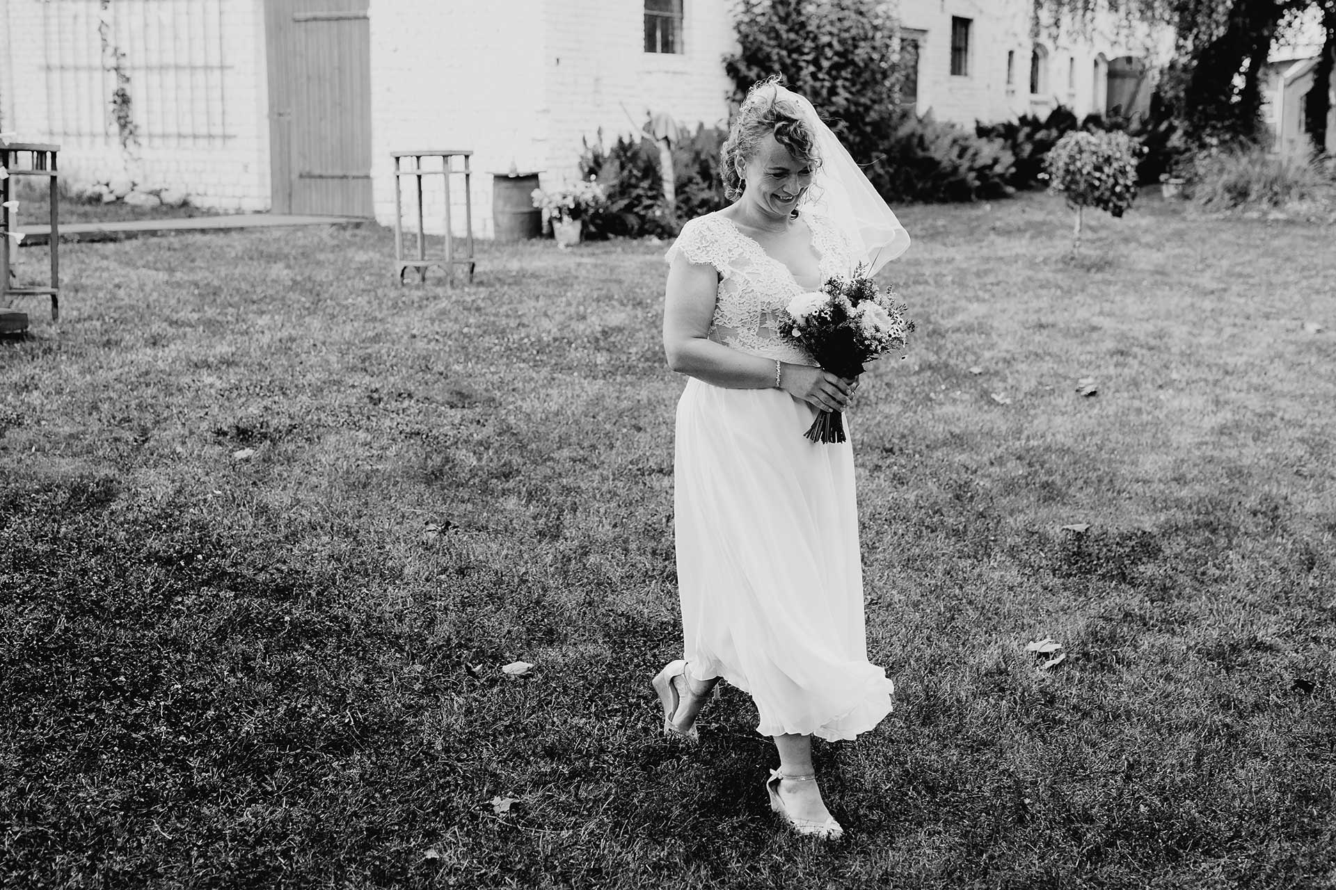 Heiraten auf dem Land: Die Braut geht mit Brautstrauß in der Hand über eine Wiese zur Trauung. Foto in schwarz-weiß vonLaura-Sophie Jung.