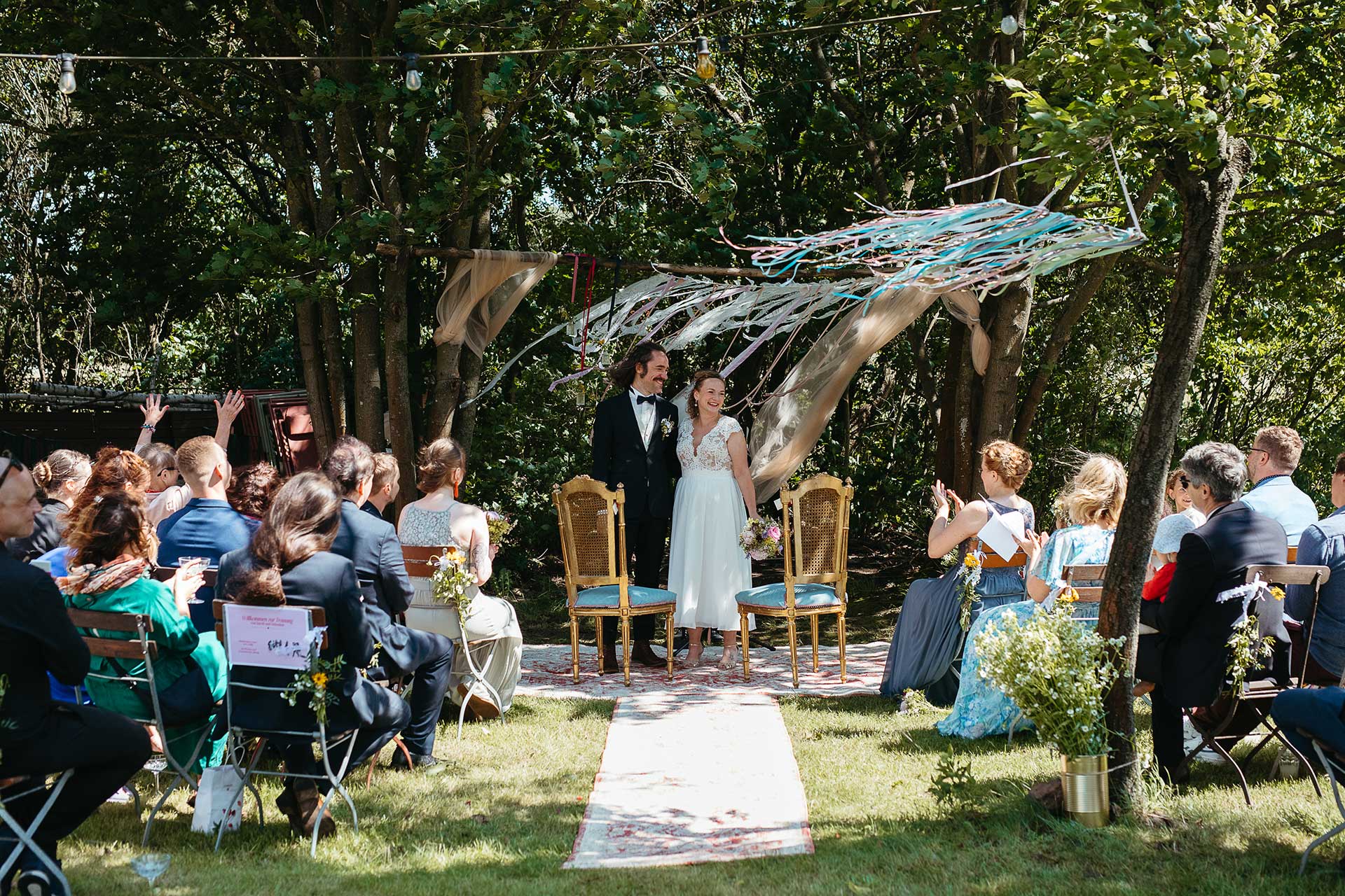 Natürliche Hochzeitsfotos auf dem Land: Brautpaar und Gäste bei der Trauung, die im Garten der Location stattfindet. Foto von Laura-Sophie Jung.