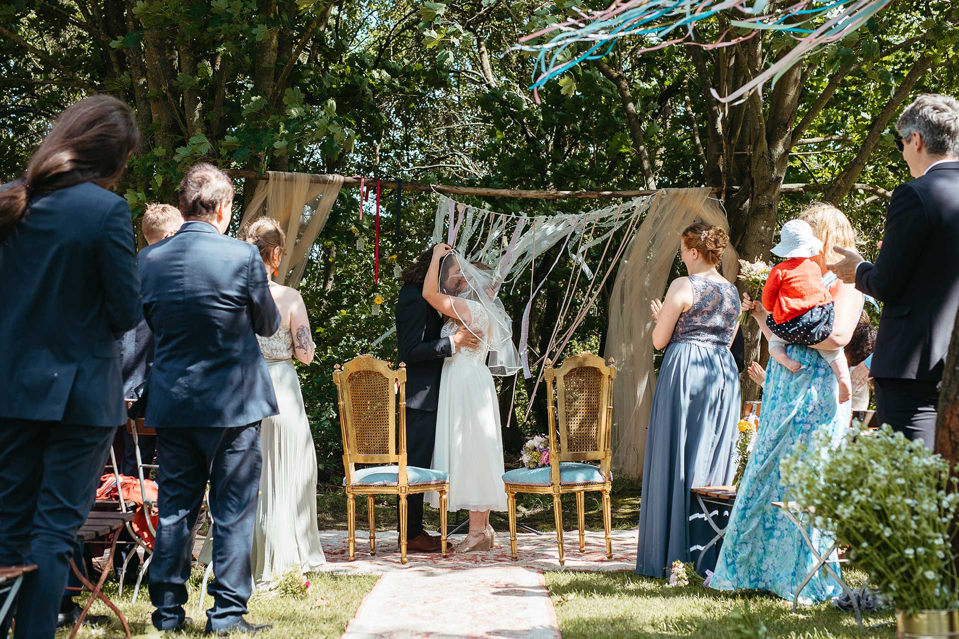 Frisch getraut: Braut und Bräutigam küssen sich, die Gäste applaudieren. Hochzeitsfoto von Laura-Sophie Jung.