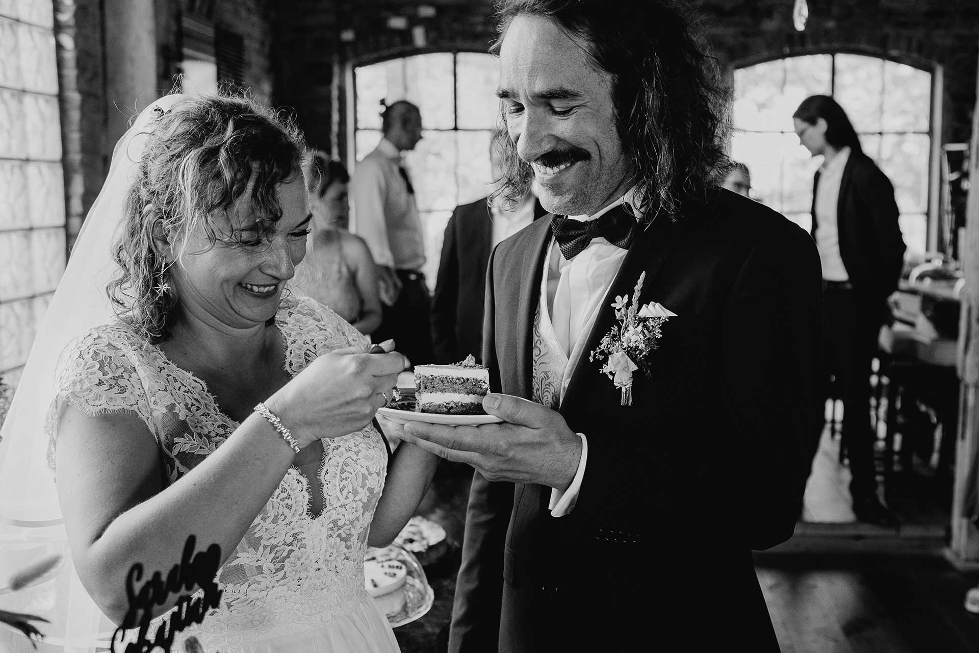 Bei der Hochzeitsfeier: Braut und Bräutigam halten gemeinsam einen Teller mit einem Stück Torte darauf, die Braut probiert. Foto in schwarz-weiß von Laura-Sophie Jung.