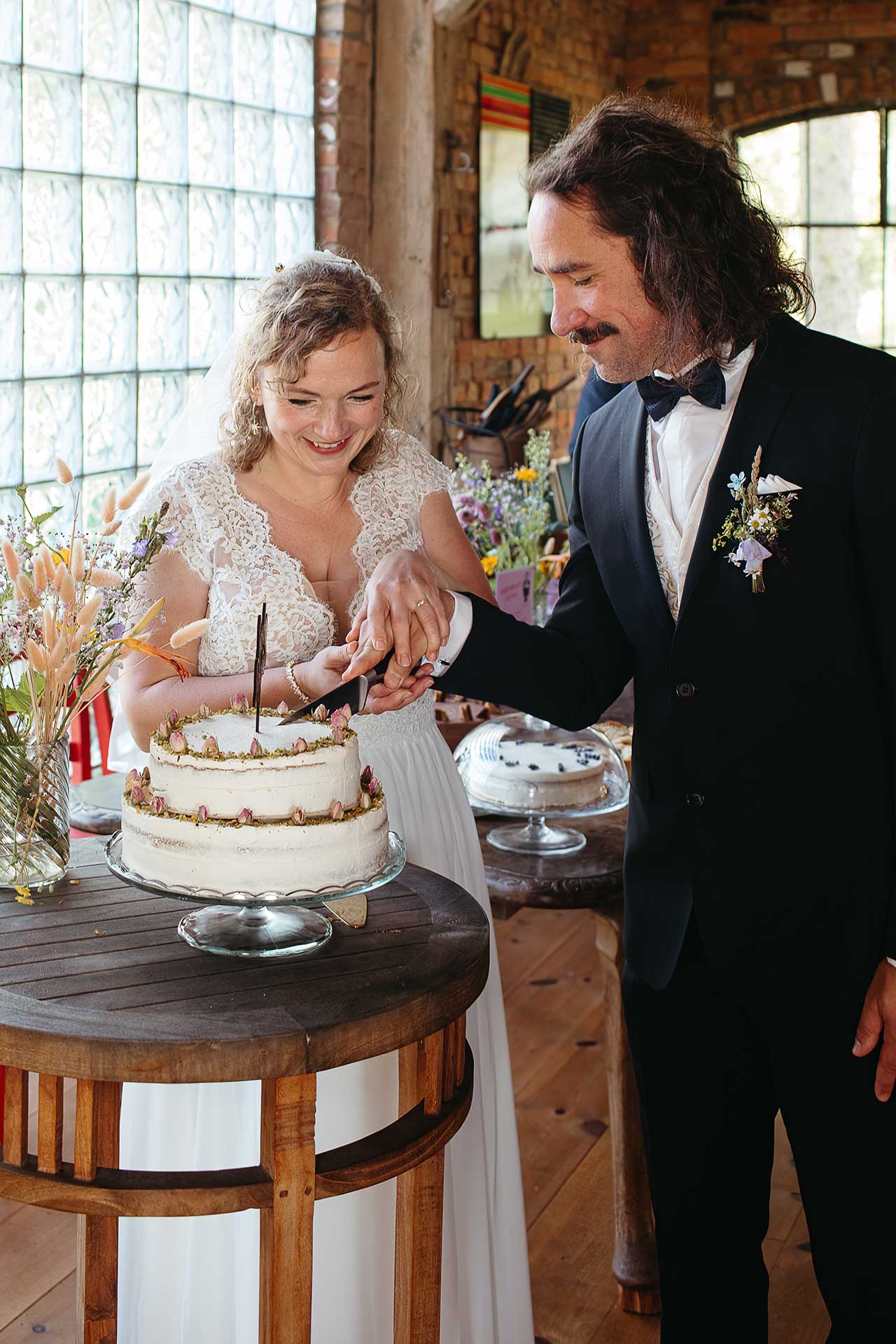 Das Brautpaar schneidet gemeinsam die Hochzeitstorte an. Im Hintegrund ein Kuchenbuffet und Dekoration mit Wiesenblumen. Hochzeitsfoto von Laura-Sophie Jung.