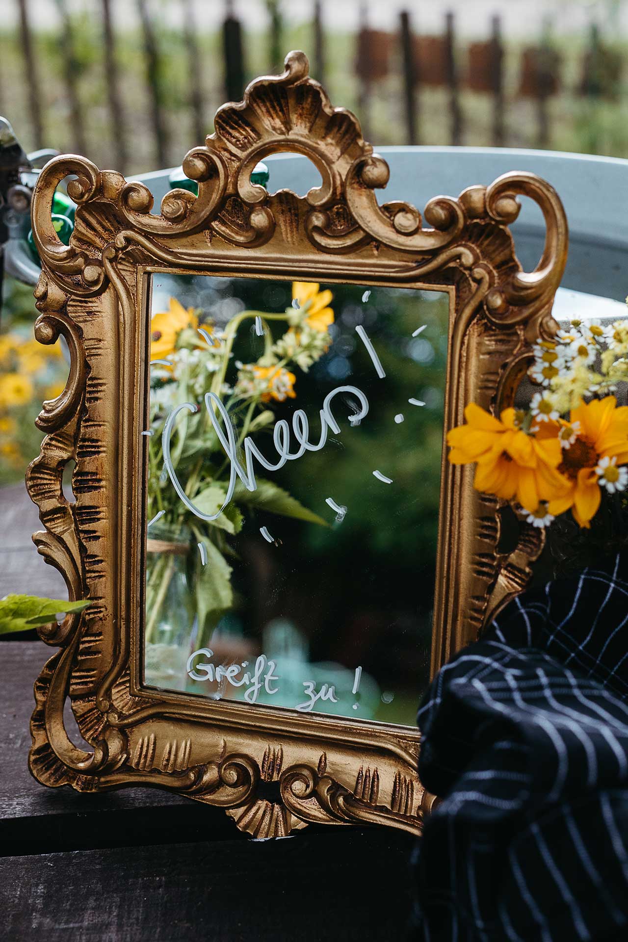 Persönlich gestaltete Hochzeitsdekoration: "Cheers" Schriftzug auf einem Spiegel mit verziertem Rahmen, im Hintergrund Blumen. Foto von Laura-Sophie Jung.