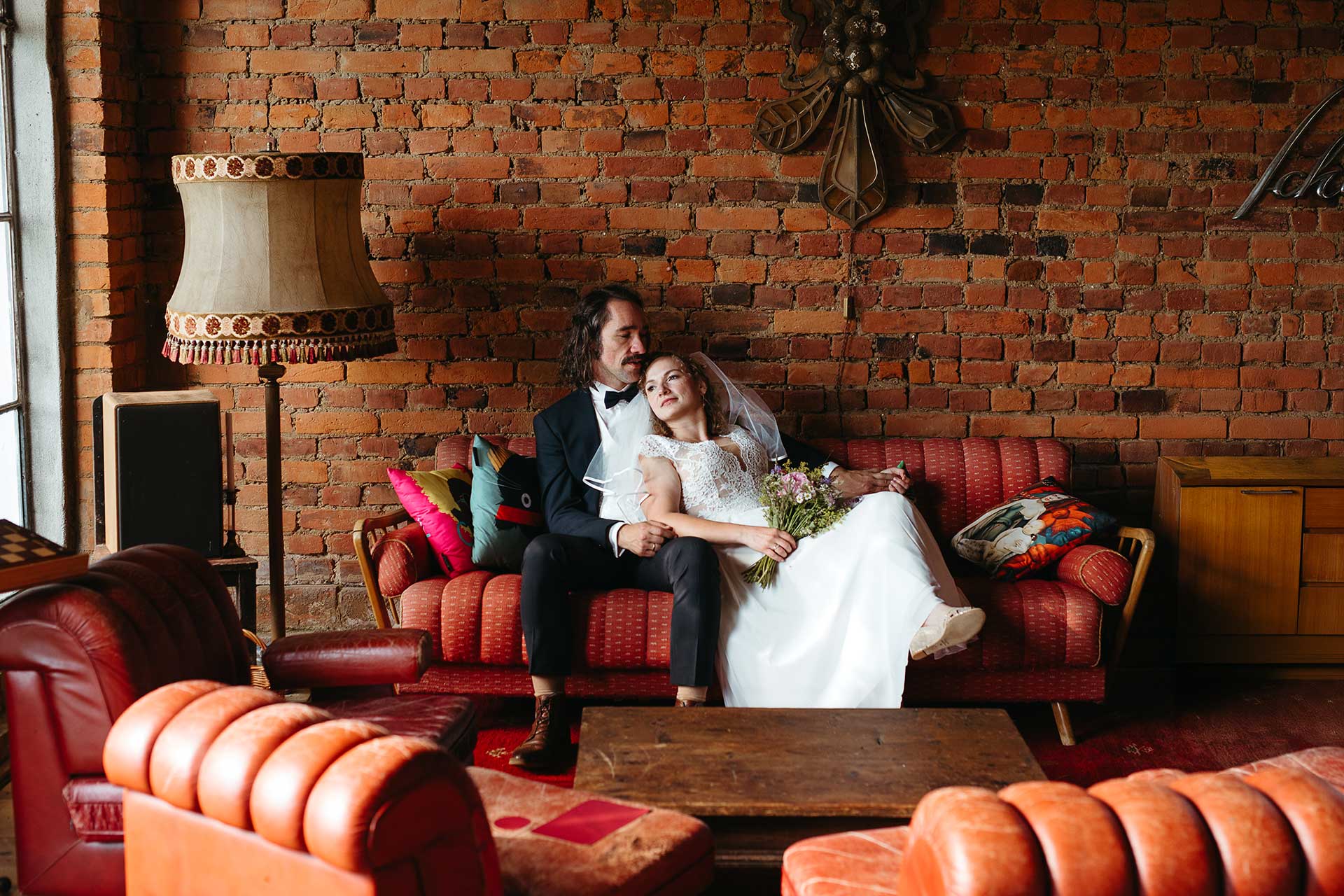 Innenbereich des "Steinreich Leben" in Brandenburg: Vintage Möbel und Ziegelwand, das Brautpaar sitzt entspannt auf einem Sofa. Foto von Laura-Sophie Jung.