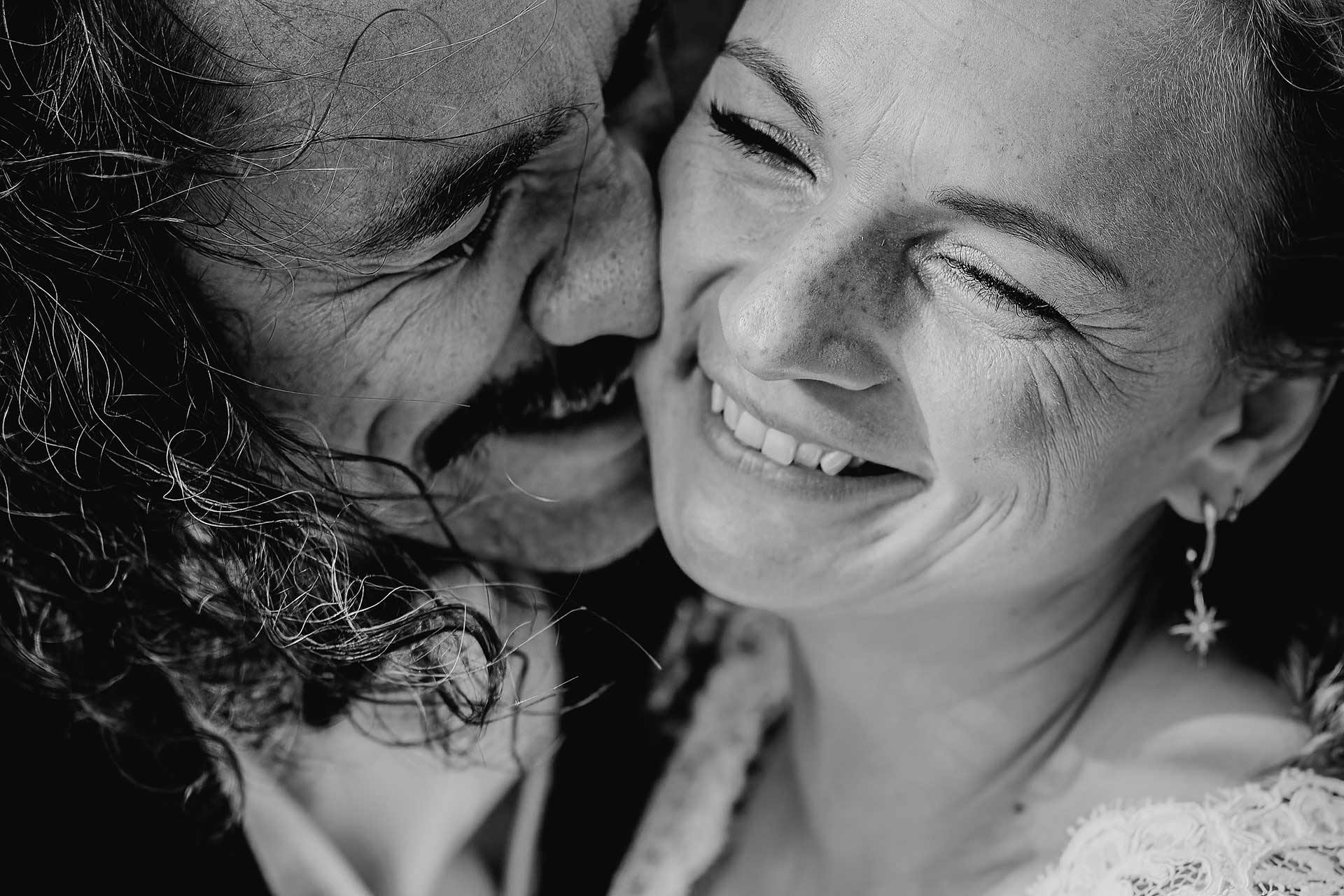 Natürliches Hochzeitsfoto in schwarz-weiß: Der Bräutigam drückt sein Gesicht überschwänglich an das Gesicht der Braut, beide lachen glücklich. Foto von Laura-Sophie Jung.