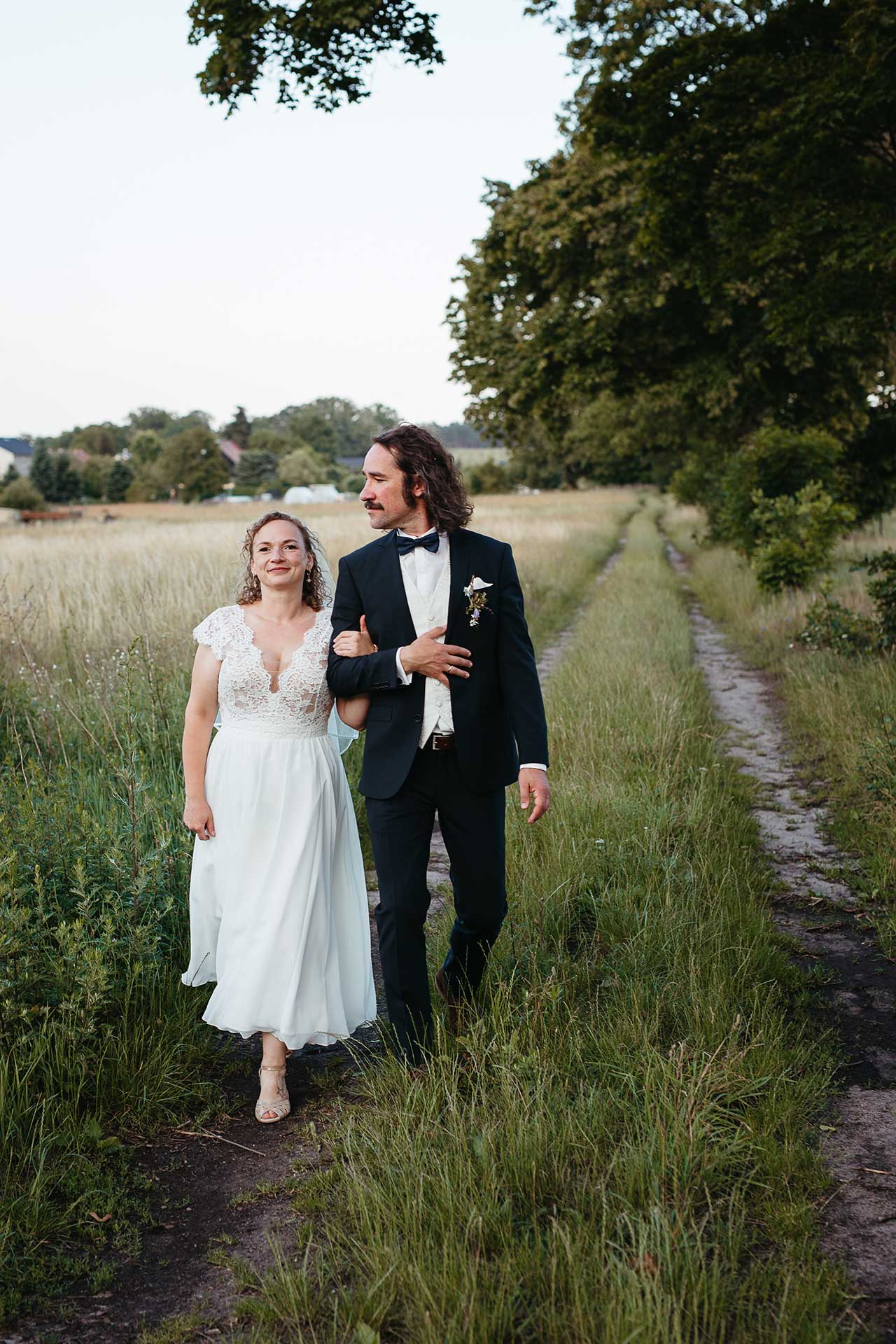 Natürliches Hochzeitsfoto mit rustikalem Charme: Das Brautpaar geht Arm in Arm auf einem Feldweg spazieren. Fotografiert von Laura-Sophie Jung.