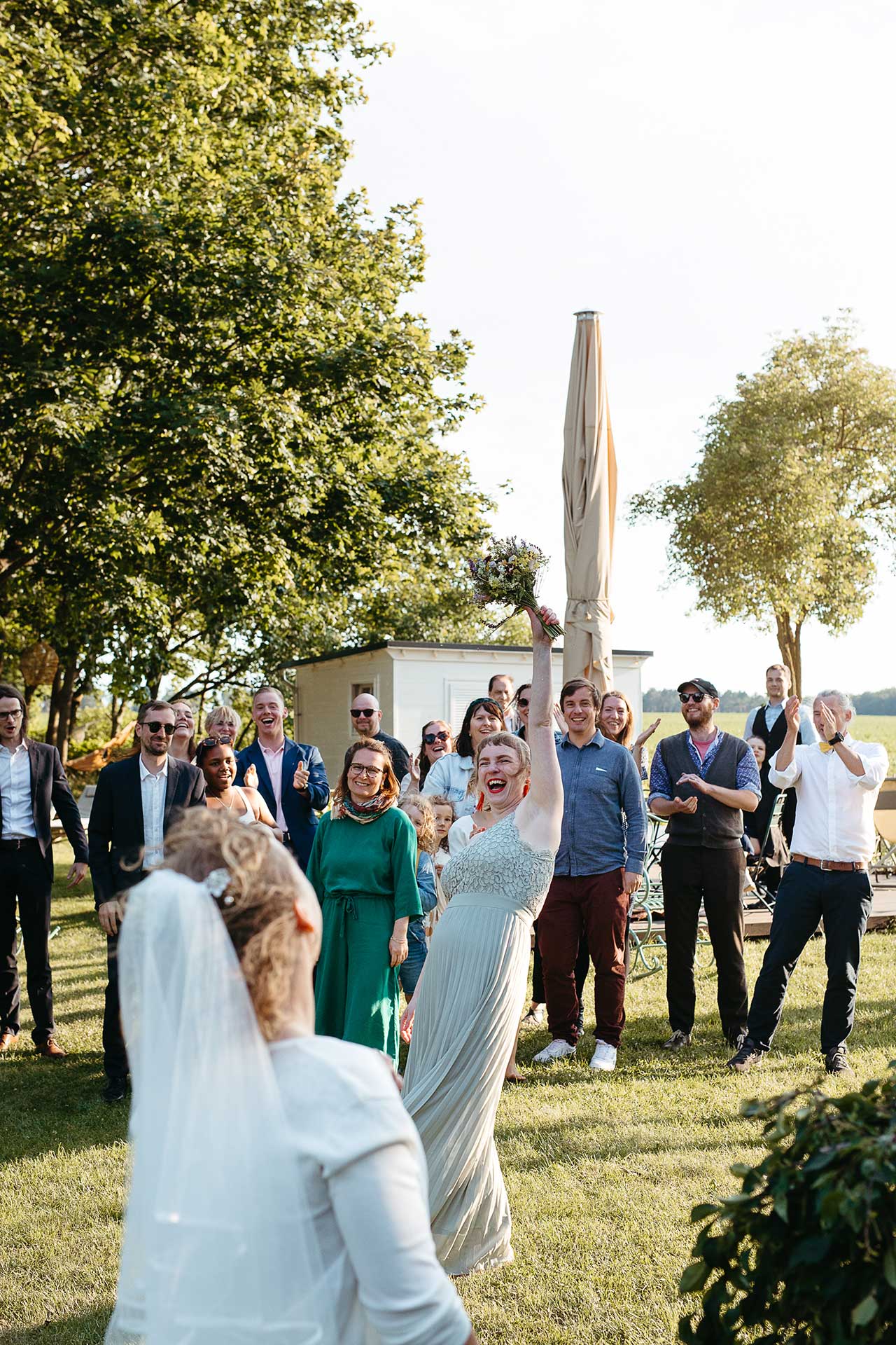 Brautstraußwurf im Garten vor der Location: Eine Frau hat den Brautstrauß gefangen und hebt ihn freudestrahlend in die Höhe. Foto von Laura-Sophie Jung.
