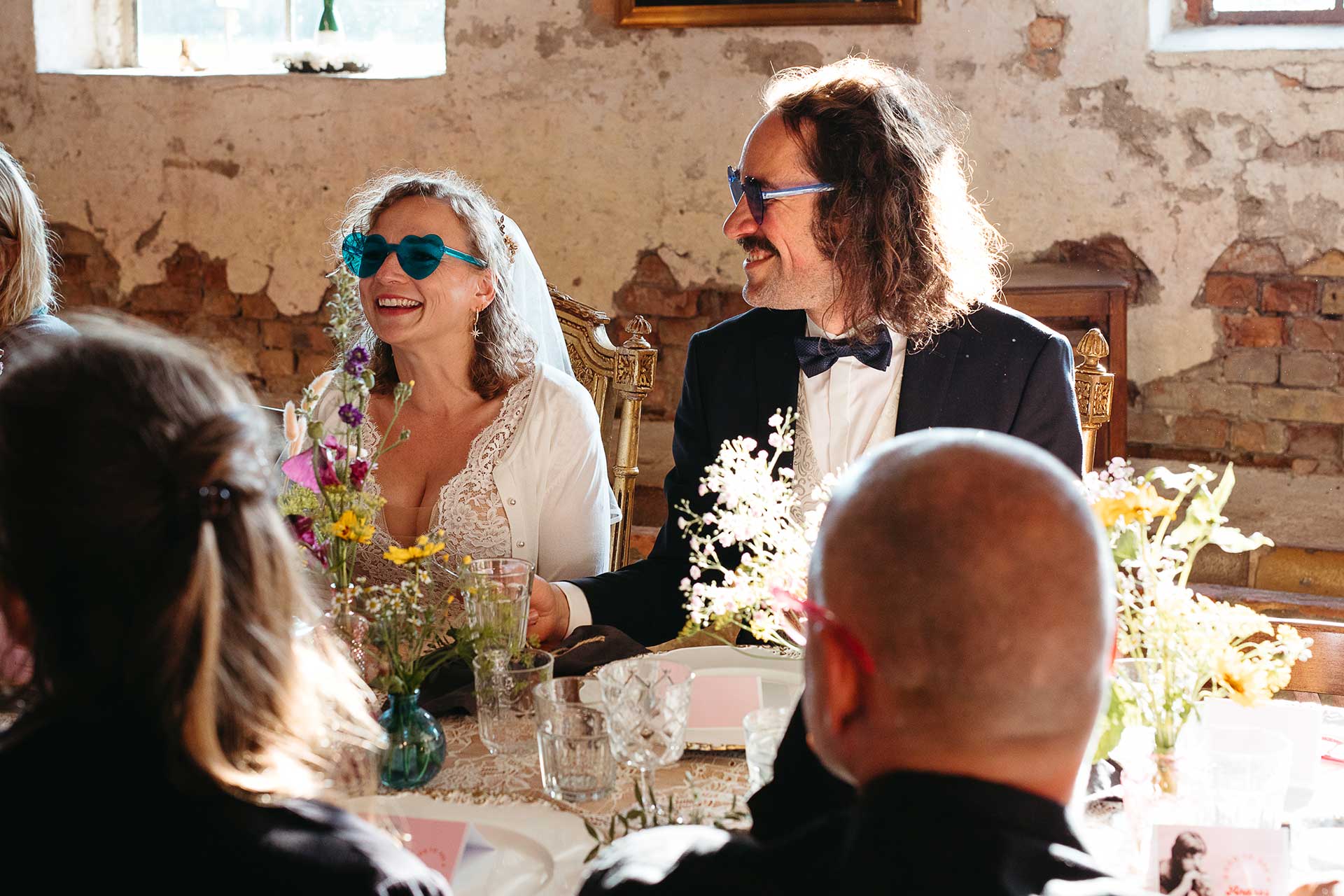 Fröhliche Hochzeitsfeier im "Steinreich Leben": Das Brautpaar sitzt mit blauen Herz-Sonnenbrillen am Tisch, lachend und scherzend. Foto von Laura-Sophie Jung.