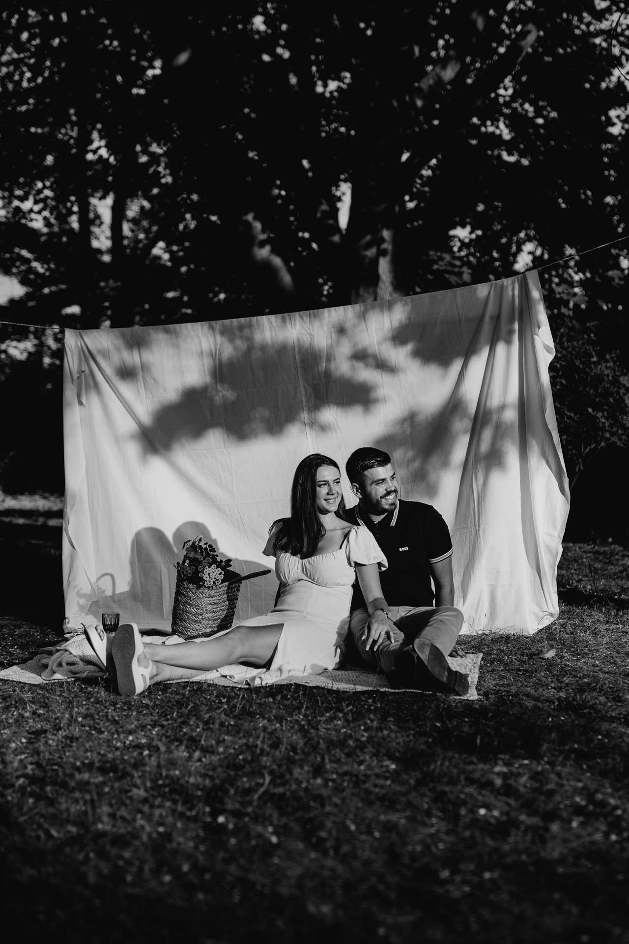 Verlobungsshooting beim Picknick Date: Paar sitzt auf einer Picknickdecke, daneben ein Picknickkorb mit Blumenstrauß. schwarz-weiß Foto von Laura-Sophie Jung, Berlin.
