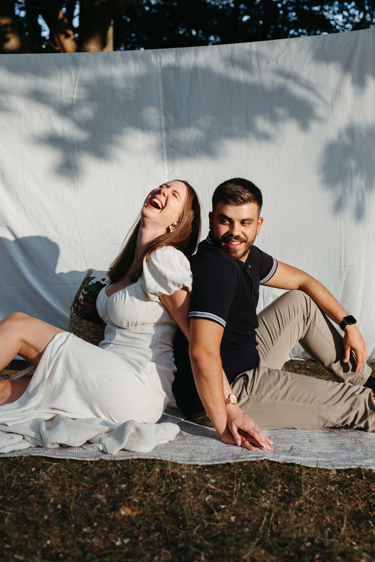 Paar beim Verlobungsshooting outdoor: Frau und Mann sitzen Rücken an Rücken, Frau lacht herzlich. Fotografiert von Laura-Sophie Jung, Berlin.