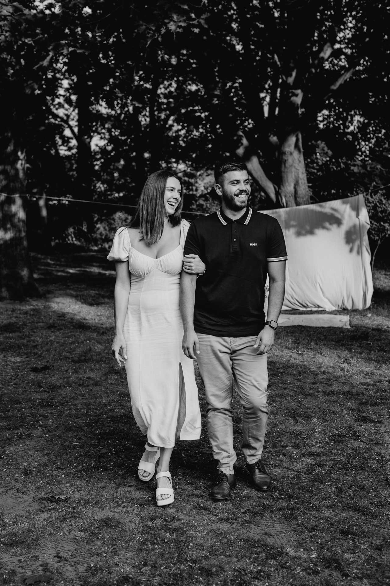 Verlobungsshooting outdoor: Paar geht Arm in Arm über eine Wiese, beide lächeln glücklich. Foto in schwarz-weiß von Laura-Sophie Jung, Berlin.