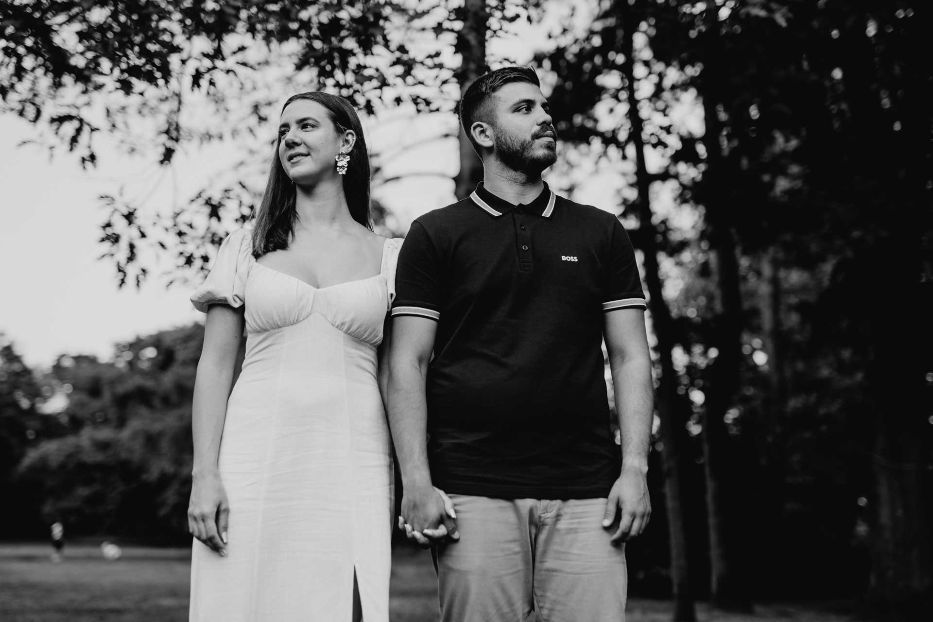 Verlobungsshooting im Park: Mann und Frau stehen Hand in Hand nebeneinander mit Blick in verschiedene Richtungen. Foto in schwarz-weiß von Laura-Sophie Jung, Berlin.